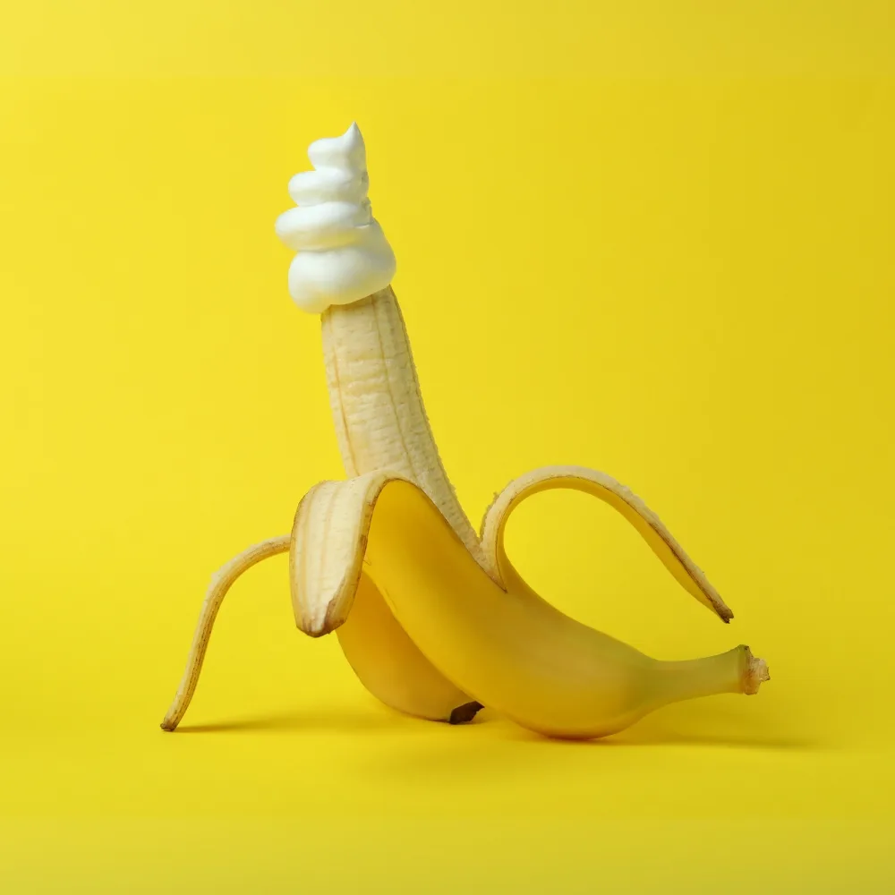 Plátano parcialmente pelado con nata en la punta sobre fondo amarillo, metáfora visual para juguetes para ellos.